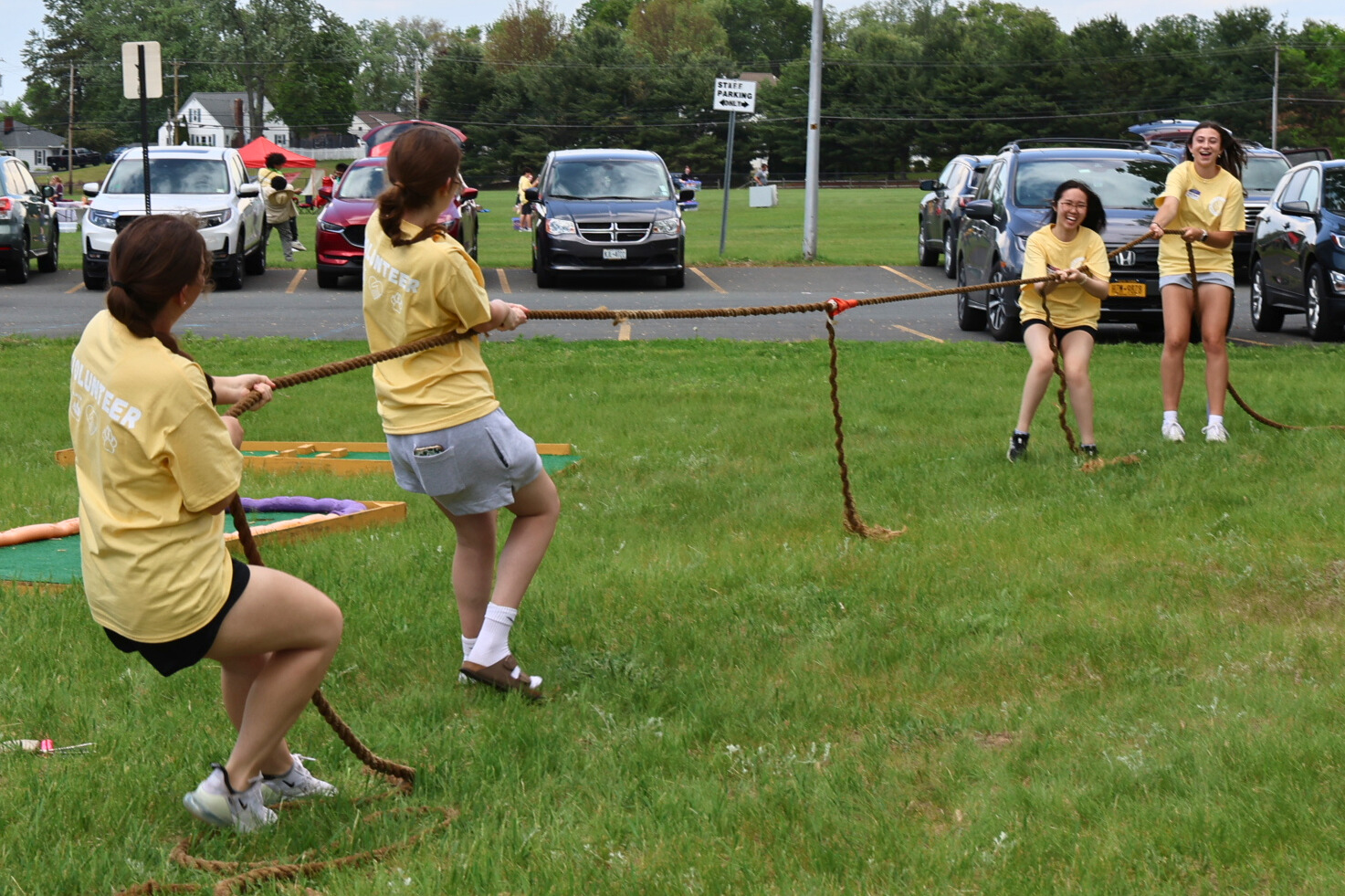 Students wearing yellow shirts play tug o' war on a grassy field in front of a parking lot